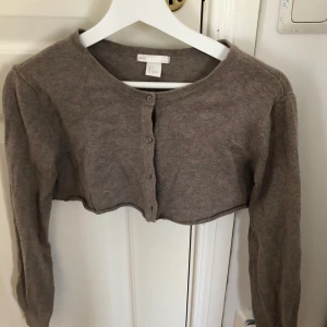 Beige croppad kofta från Basic - Säljer en beige croppad kofta från Basic. Den har långa ärmar och knappar framtill. Perfekt för lager-på-lager-stil eller som ett lättare plagg för svalare dagar.