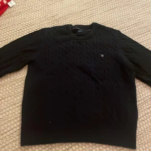 Mörkblå tröja från Gant - Säljer en stilren svart stickad tröja från Gant med ett diskret mönster. Tröjan har långa ärmar och en rund halsringning. Perfekt för kyligare dagar.