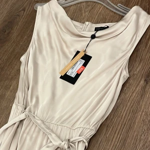 Champagnefärgad klänning från Stockh LM - Elegant champagnefärgad klänning från Stockh LM med ett knytband i midjan och dragkedja bak. Klänningen har en mjuk, glansig yta och är ärmlös med en draperad halslinje. Knälång! 