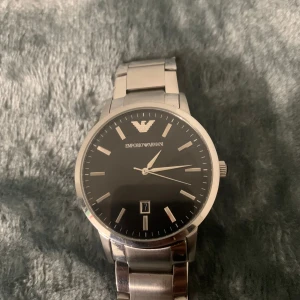Silverfärgad klocka från Emporio Armani - Stilren klocka från Emporio Armani med svart urtavla och datumvisning. Klockan har en silverfärgad länkarmband och det ikoniska logotypen på urtavlan. Perfekt för en elegant look. 