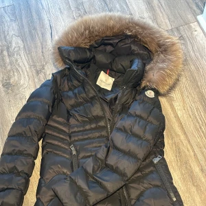  Moncler jacka  - (Äkta)Snygg svart dunjacka från Moncler med en lyxig pälsdetalj på huvan. Jackan har en figursydd passform och är utrustad med dragkedjor framtill och på fickorna. Runt storlek 36 passar både mindre och större!