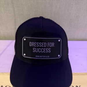 Svart keps från John Hatter & Co - Snygg svart keps med texten 'Dressed for Success' på framsidan. Märket John Hatter & Co är känt för sina stilrena och moderna accessoarer. Perfekt för att ge din outfit en cool touch.