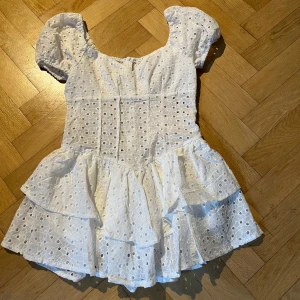 Vit klänning med volanger - Charmig vit klänning med korta puffärmar och volanger. Klänningen har ett broderat mönster och en söt, feminin stil. Perfekt för somriga dagar. Ngt liten i storleken. Helt ny enbart provad. 