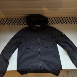 Svart jacka med huva - Snygg svart jacka från Stone island med huva och dragkedja framtill. Jackan har två bröstfickor med knappar och en stilren design. Perfekt för en casual look.