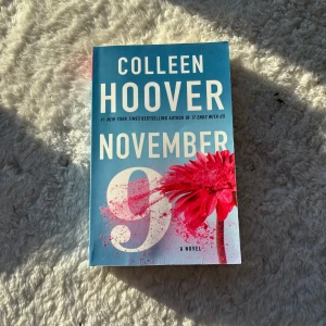 November 9 - Boken 'November 9' av Colleen Hoover är en gripande roman om kärlek och öde. Omslaget är blått med en stor rosa blomma och vit text. Perfekt för dig som älskar romantiska berättelser med oväntade vändningar.