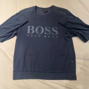 Blå tröja från Hugo Boss - Säljer en stilren blå tröja från Hugo Boss med deras logga tryckt på framsidan. Tröjan har en rund halsringning och ribbade muddar vid ärmslut och nederkant. Perfekt för en avslappnad men snygg look.