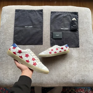 Vita sneakers med röda stjärnor från Saint Laurent - Snygga vita sneakers från Saint Laurent med röda stjärnor och blå detaljer på hälen. Skorna har en klassisk design med snörning och en bekväm sula. Perfekta för att ge din outfit en unik touch.