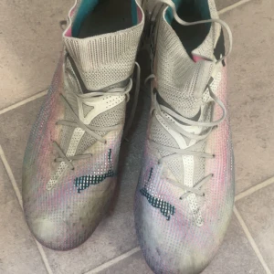 Flerfärgade fotbollsskor från Puma - Säljer ett par coola fotbollsskor från Puma i flerfärgat mönster med inslag av rosa, blått och grått. Skorna har snörning och en stickad överdel för bra passform. Sulan är rosa med dobbar för optimalt grepp på planen.