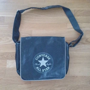 Svart axelväska från Converse - Säljer en svart axelväska från Converse med det klassiska Chuck Taylor All Star-logotypen på framsidan. Väskan har ett justerbart axelband och flera fack för praktisk förvaring. Perfekt för vardagsbruk eller skola.