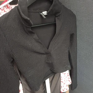 Svart ribbad tröja från Divided - Säljer en svart ribbad tröja från Divided med en unik design. Tröjan har en v-ringad stil och långa ärmar, perfekt för en stilren look. Passar bra till både jeans och kjol. Perfekt för höst och vinter!