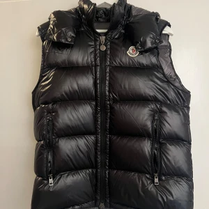 Svart dunväst från Moncler - Snygg svart dunväst från Moncler med dragkedja och hög krage. Västen har två praktiska fickor med dragkedjor och Monclers logga på bröstet. Perfekt för kyliga dagar när du vill hålla stilen.