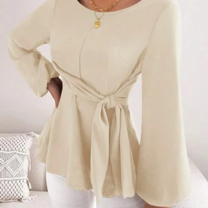 Beige blus med knytband - Elegant beige blus med långa ärmar och ett knytband i midjan för en smickrande passform. Blusen har en rund hals och vida ärmar som ger en luftig känsla. Perfekt för en stilren look.