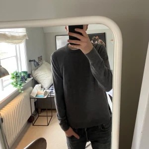 Grå tröja från These Glory Days - Säljer en stilren grå tröja från These Glory Days, tillverkad i mjuk merinoull. Perfekt för kyligare dagar med sin klassiska design och långa ärmar. Passar både till jeans och mer uppklädda tillfällen.