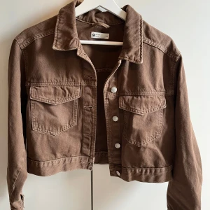 Brun croppad jacka från Gina Tricot - Snygg brun croppad jacka från Gina Tricot med knappar framtill och två bröstfickor. Perfekt för en avslappnad stil. Jackan har långa ärmar och en klassisk krage.