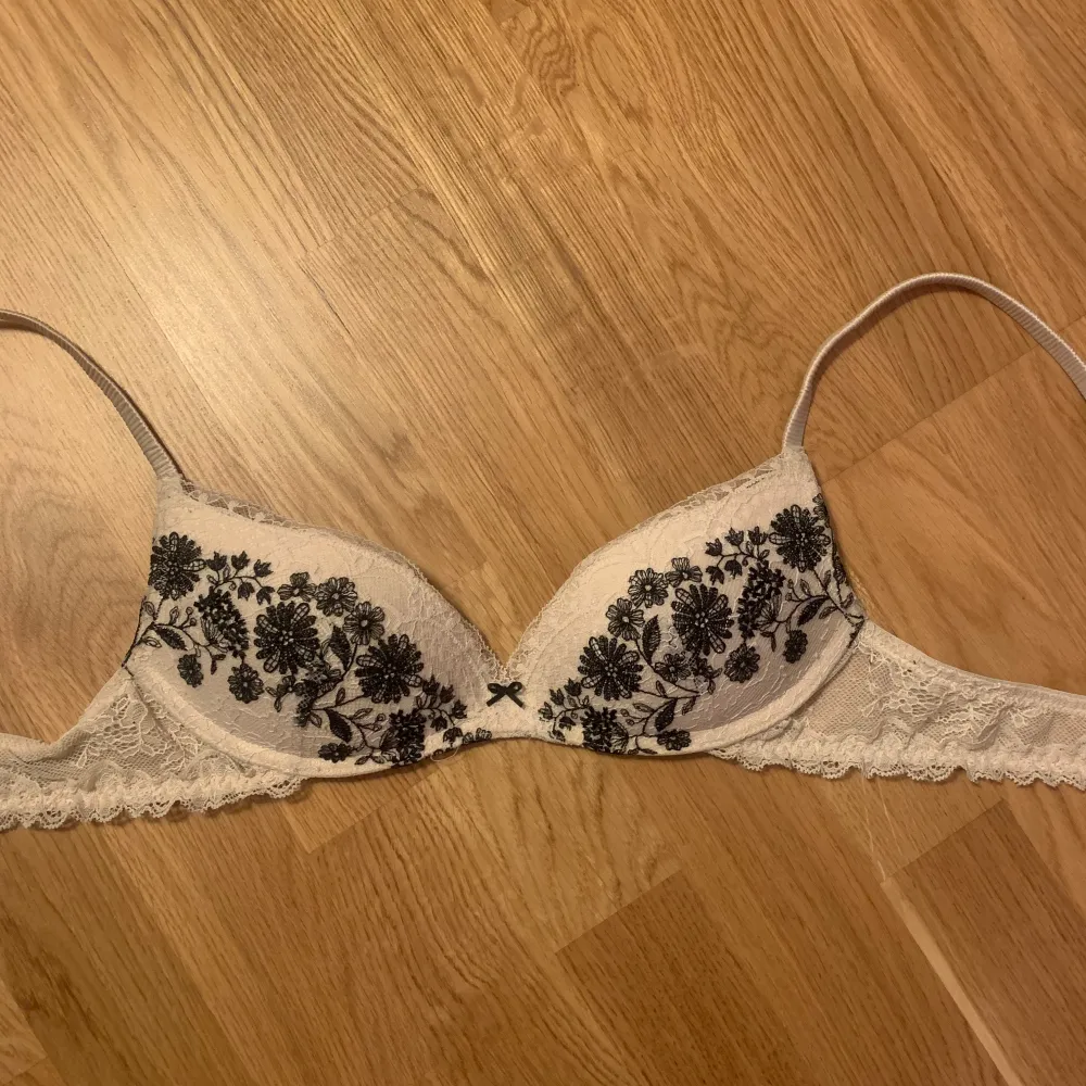 Säljer en elegant vit bh med svarta blommiga broderier och spetsdetaljer. Bh:n har justerbara axelband och en liten rosett framtill. Perfekt för att känna sig extra fin.. Muu.