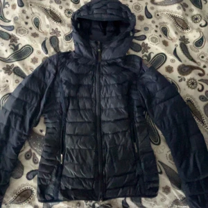 Mörkblå dunjacka från Parajumpers - Säljer en mörkblå dunjacka från Parajumpers. Jackan är superlätt och har en dragkedja framtill samt en huva. Perfekt för kyliga dagar och har en snygg logga på ärmen. Passar dig som vill ha en stilren och funktionell jacka.