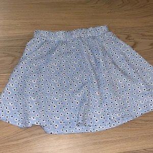 Blå blommig kjol - Söt blå kortkjol med ett blommigt mönster i vitt och svart. Kjolen har en elastisk midja för en bekväm passform. Perfekt för varma dagar och ger en fräsch look. 