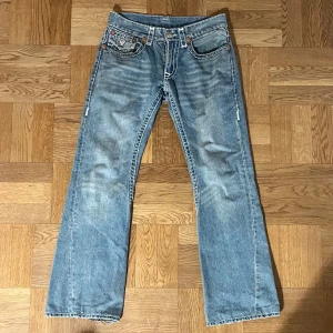 Blå jeans från True Religion - Säljer ett par sköna blå jeans från True Religion med snygga kontrastsömmar och ikoniska bakfickor. Modellen är Joey Super T med en bootcut passform och låg midja. 