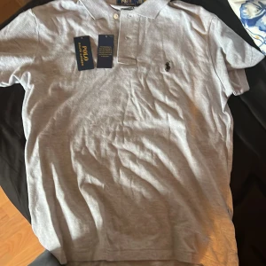Grå pikétröja från Polo Ralph Lauren - Säljer en stilren grå pikétröja från Polo Ralph Lauren. Tröjan har korta ärmar och en klassisk krage med knappar. Den ikoniska loggan är broderad på bröstet. Perfekt för en avslappnad men ändå snygg look. Perfekt nu när det blir sommar. Säljer den eftersom den är inte riktigt min stil längre och har inte kommit till så mycket användning. Den är storlek L MEN PASSAR M OCKSÅ