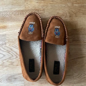 Bruna mockaloafers från Polo Ralph Lauren - Säljer ett par bruna mockaloafers från Polo Ralph Lauren med broderad björn på ovansidan. Skorna har en mysig foder inuti och en klassisk design med synlig söm runt kanten. Perfekta för att hålla fötterna varma och bekväma.