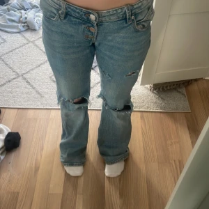 Blå slitna jeans - Säljer ett par blå jeans med slitningar och knappgylf. De har en avslappnad passform och är perfekta för en casual look. Jeansen har en klassisk femficksdesign och är tillverkade i denim.