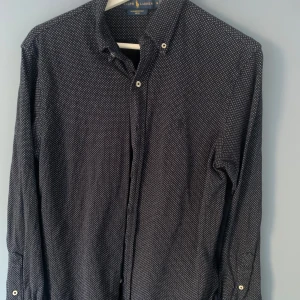 Svart prickig skjorta från Ralph Lauren - Säljer en stilren svart skjorta från Ralph Lauren med små vita prickar. Skjortan har långa ärmar och knappar framtill. Perfekt för en elegant look.