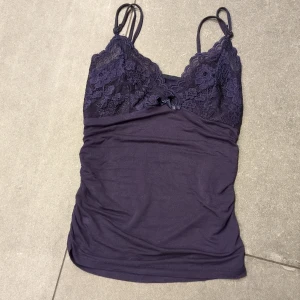 Mörklila linne med spetsdetaljer - Säljer ett mörklila linne med vackra spetsdetaljer och smala axelband. Linnet har en liten rosett framtill och är perfekt för en stilren look.