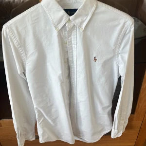 Vit skjorta från Ralph Lauren - Säljer en klassisk vit skjorta från Ralph Lauren i slim fit. Skjortan har långa ärmar och knappar framtill samt en broderad logotyp på bröstet. Tröjan är använda ett fåtal gånger och i bra skick cirka 8,5/10. Säljer varan eftersom den inte passar längre. 