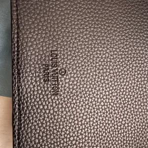 Brun axelväska  - Säljer en snygg brun axelväska med det klassiska monogrammönstret. Väskan har en justerbar rem och en praktisk ficka framtill. Perfekt för att bära med sig det viktigaste med stil.