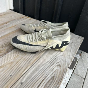 Nike fotbollsskor i beige och svart - Snygga fotbollsskor från Nike i en stilren beige färg med svarta detaljer och snörning. Perfekta för fotbollsplanen med en tight passform och slitstark design.