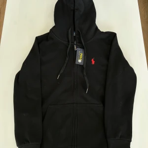 Svart Ralph Lauren Zip Up - Säljer en stilren svart hoodie från Ralph Lauren med dragkedja och det klassiska röda logotypen på bröstet. Perfekt för en avslappnad stil med justerbar huva och ribbade muddar.