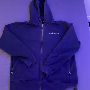 Blå hoodie från Sail Racing - Säljer en snygg blå hoodie från Sail Racing med dragkedja och huva. Perfekt för kyliga dagar. Den har en stilren logga på bröstet och praktiska fickor. Passar både till vardags och mer avslappnade tillfällen.