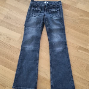 MörkGrå jeans från Gina Tricot - Snygga grå jeans från Gina Tricot med bootcut-stil. Byxorna har dekorativa fickor med knappar framtill och klassiska bakfickor. Perfekta för en avslappnad look.