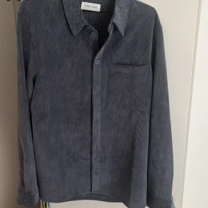 Grå manchesterskjorta från Pier One - Snygg grå manchesterskjorta från Pier One med klassisk krage, bröstficka och knäppning framtill. Skjortan har lång ärm och är tillverkad i mjuk manchester som ger en cool och avslappnad vibe. Perfekt att bära öppen över en t-shirt eller stängd för en stilren look. Nypris 200 säljer för 70