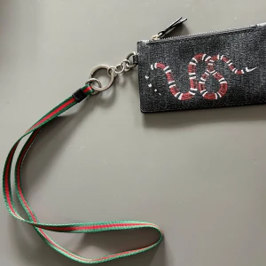 Gucci svart plånbok med orm - Svart plånbok från Gucci i skinn med ikonisk röd och vit orm på framsidan. Har dragkedja upptill, flera kortfack på baksidan och en grön-röd axelrem med metallringar. Snygg och edgy accessoar med lyxig känsla.