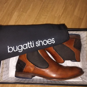 Bruna Chelsea boots från Bugatti - Snygga Chelsea boots från Bugatti i brunt skinn med svarta elastiska paneler på sidorna och diskret mönstrad detalj vid hälen. Skorna har låg klack, rund tå och klassisk platt sula. Perfekta för dig som vill ha en stilren och tidlös look. Nypris: 1350kr