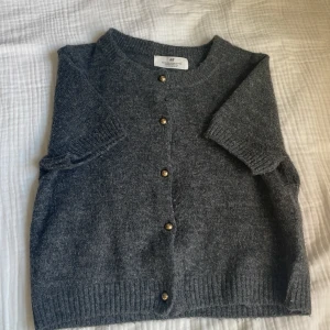 Grå stickad kofta från H&M - Kortärmad grå kofta från H&M med rund hals och snygga guldfärgade knappar framtill. Mjuk stickad känsla och ribbade muddar vid ärmslut och nederkant. Perfekt för lager-på-lager och snygg stil. Orginalpris ca 200kr. Pris kan diskuteras.