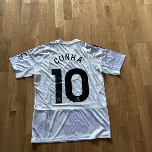Vit Manchester United Cunha #10 tröja - Säljer en vit Manchester United fotbollströja med Cunha och nummer 10 på ryggen. Tröjan har svarta detaljer, Adidas-logga, klubbmärke och Snapdragon sponsortryck på bröstet. Klassisk V-ringning och svarta ränder på axlarna. Tillverkad i lätt polyester.