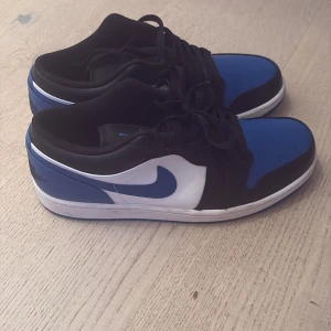 Nike Air Jordan 1 Low blå och svart - Nike Air Jordan 1 Low sneakers med en snygg färgkombo i blått, svart och vitt. Skorna har klassisk snörning, perforerad tå och den ikoniska swoosh-loggan på sidan. Materialet är skinn och syntet, med rund tå och platt sula. Perfekt för dig som vill ha en stilren och sportig look. Skorna är knappt använda. Öppen för prisförslag. 