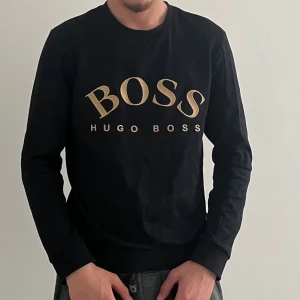 Svart sweatshirt från Hugo Boss - Svart sweatshirt från Hugo Boss med stort guldigt BOSS-tryck på bröstet. Klassisk rund halsringning och långa ärmar. Tröjan är i mjuk bomull och har en clean, stilren look som passar perfekt till jeans eller joggers.