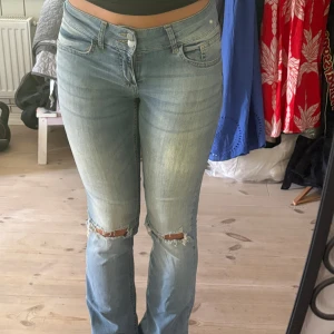 Ljusblå bootcut jeans med slitna knän - Säljer ett par ljusblå bootcut jeans med snygga slitningar över båda knäna. Jeansen har klassisk femficksdesign, hällor för bälte och en lätt tvättad look. Materialet är mjukt denim och passformen är normal med lite utsvängda ben.