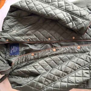 Quiltad grön jacka från U.S. Polo Assn. - Helt ny snygg quiltad jacka i mörkgrönt från U.S. Polo Assn. Jackan har tryckknappar, dragkedja och brunt kantband. 