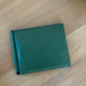 R0lex plånbok - Snygg grön plånbok i skinn med stilrent mönster och flera kortfack på insidan. Plånboken har en metallklämma för sedlar och en smidig, kompakt design som passar perfekt i fickan. Perfekt för dig som vill ha koll på kort och cash med stil. Nypris: 3000kr