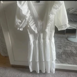 Vit volangklänning från Gina Tricot - Supersöt vit kortklänning från Gina Tricot med v-ringning, volanger längs halsringningen och puffiga kortärmar. Klänningen har smock i midjan och ett lätt, krinklat tyg som ger en luftig känsla. Perfekt för sommarens alla tillfällen!