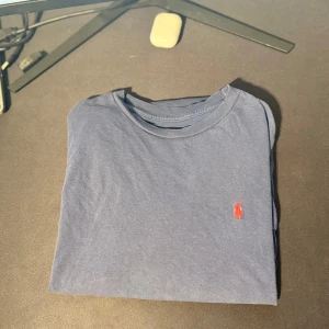 Blå t-shirt från Polo Ralph Lauren - Snygg marinblå t-shirt från Polo Ralph Lauren med klassisk röd logobrodyr på bröstet. T-shirten har rund hals och är kortärmad, tillverkad i mjuk bomull som är skön mot huden. Perfekt för dig som gillar stilrena och ikoniska plagg.