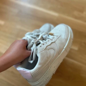 Nike Air Force 1 Pixel i beige mocka - Nike Air Force 1 Pixel sneakers i beige mocka med chunky vit plattformssula och ljusrosa detaljer på hälen. Klassisk snörning med guldfärgad detalj på skosnöret. Snyggt broderad swoosh på sidan och coola pixel-inspirerade detaljer på sulan. Storlek 40, används ca 2 ggr