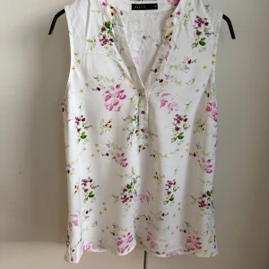 Vit blommig ärmlös blus från Mohito - Supersöt vit blus från Mohito med blommigt mönster i rosa, lila, grönt och gult. Blusen är ärmlös och har en v-ringad hals med tre små knappar framtill. Perfekt för dig som gillar fräscha och somriga vibes.