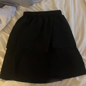 Svart volangkjol  - Snygg svart kjol med volangdetaljer och resår i midjan. Kjolen har en enkel, stilren look och är lätt att matcha med olika toppar. Passformen är normal och kjolen slutar ovanför knäna. Perfekt för dig som gillar klassisk stil med en twist.