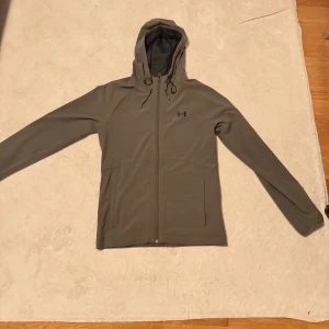 Under Armour grön vindjacka med huva - Snygg grå vindjacka från Under Armour med huva och dragsko. Jackan har dragkedja framtill, två fickor och diskret logga på bröstet. Perfekt för blåsiga dagar och sportiga outfits. Lätt och smidig att bära med sig.