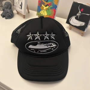 Corteiz Keps - Snygg svart keps med mesh på sidorna och justerbar snapback. Framtill finns broderade vita stjärnor och ett motiv i silver. Märkt med Corteiz på insidan. Perfekt för dig som gillar streetstyle och vill sticka ut med coola detaljer.